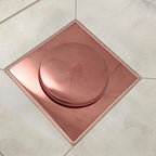 Ralo click inteligente 10x10 cm rose gold instalado em piso de porcelanato claro, com tampa redonda