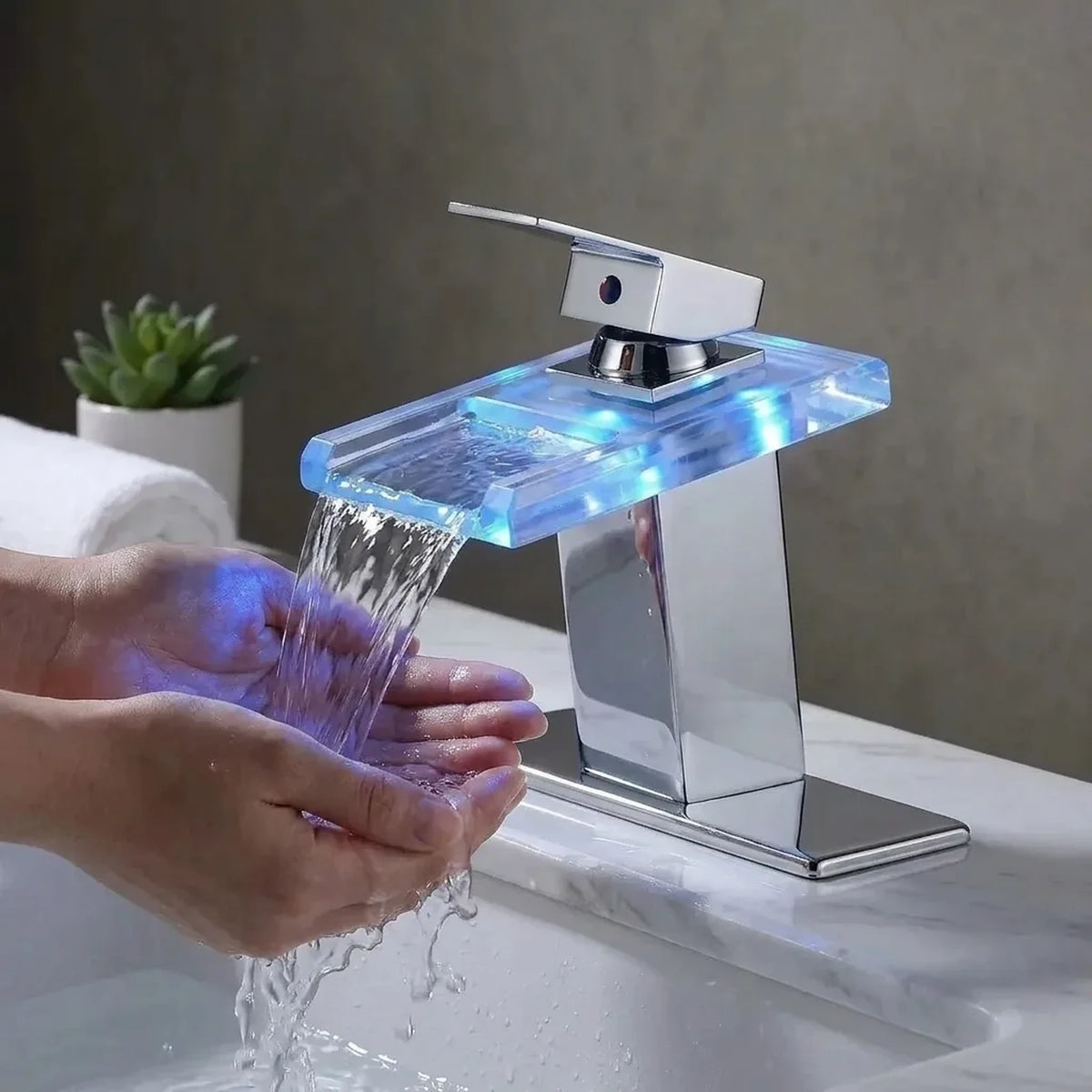 Torneira AquaGlam cromada em uso: mãos sob a lâmina de água em cascata com LED azul, na cuba do banheiro